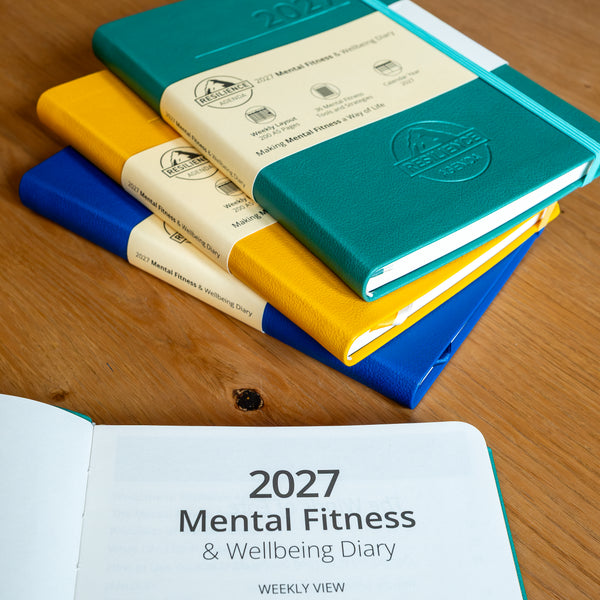 A stack of hardcover notebooks on a wooden tabletop, with one teal notebook on top showing a cream paper band and embossed logo. An open view at the bottom shows the cover text '2027 Mental Fitness & Wellbeing Diary' on a teal cover.