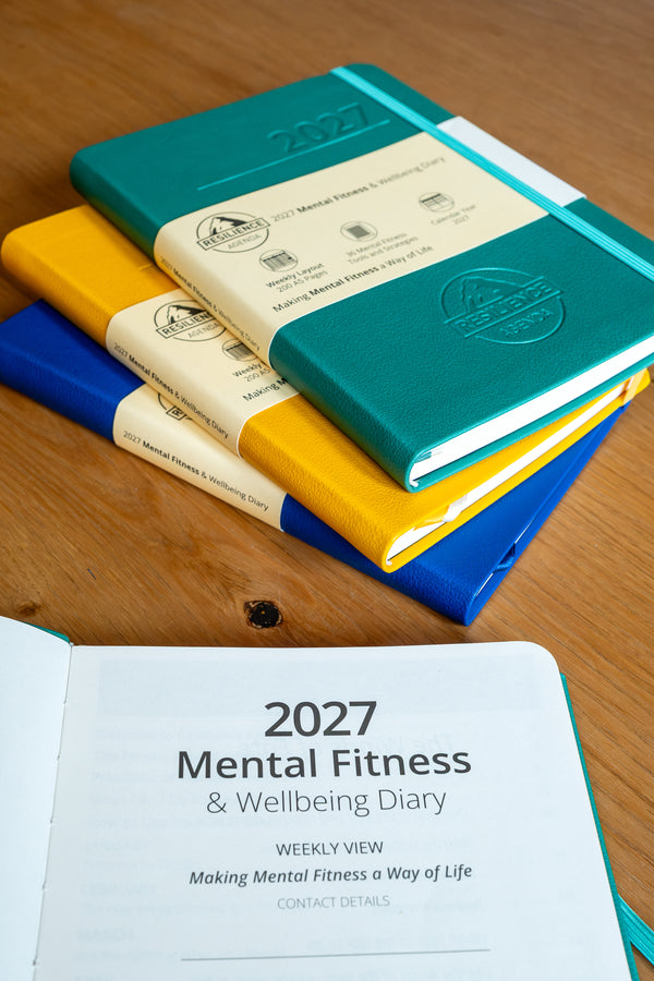 A stack of hardcover notebooks on a wooden tabletop, with one teal notebook on top showing a cream paper band and embossed logo. An open view at the bottom shows the cover text '2027 Mental Fitness & Wellbeing Diary' on a teal cover.