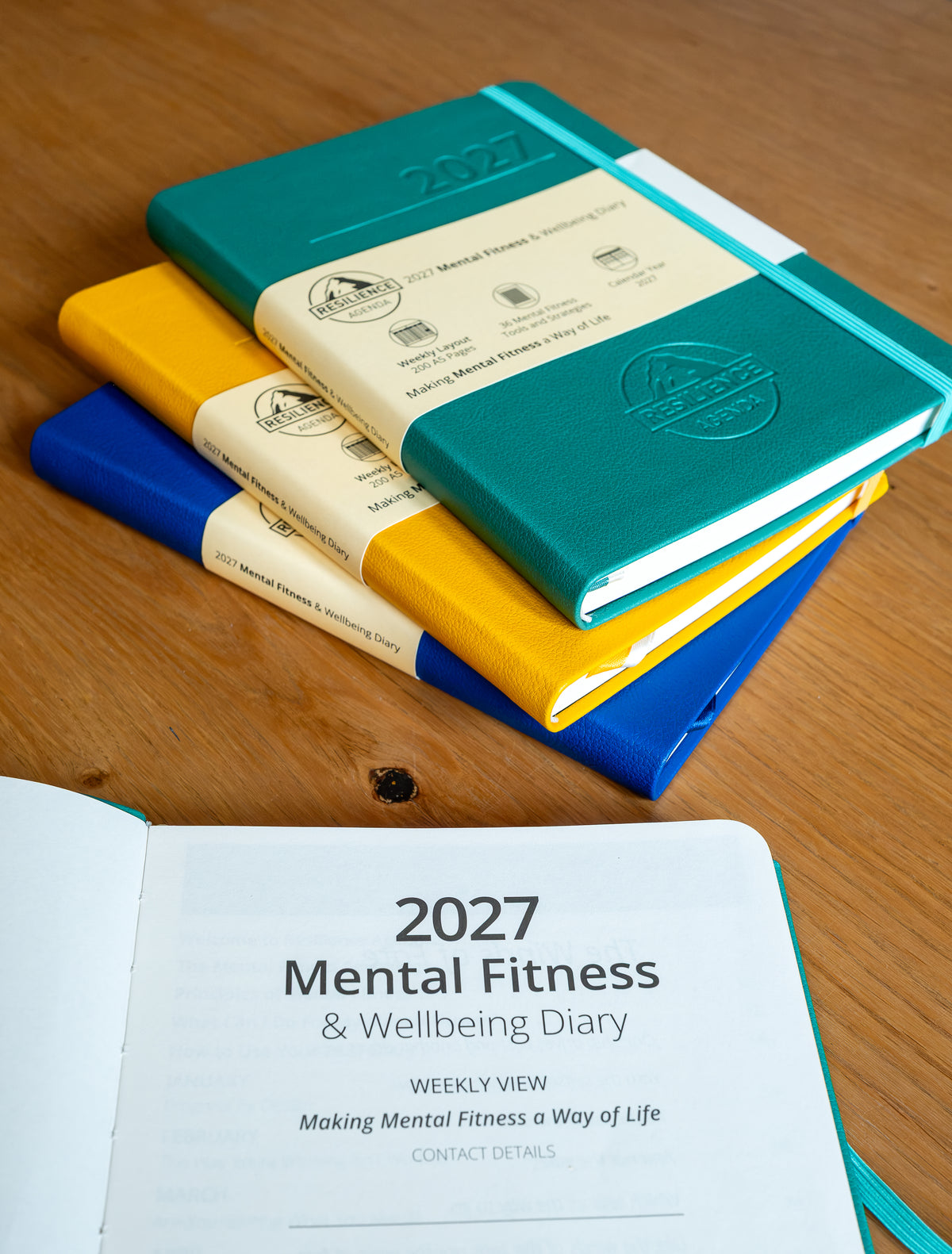 A stack of hardcover notebooks on a wooden tabletop, with one teal notebook on top showing a cream paper band and embossed logo. An open view at the bottom shows the cover text '2027 Mental Fitness & Wellbeing Diary' on a teal cover.